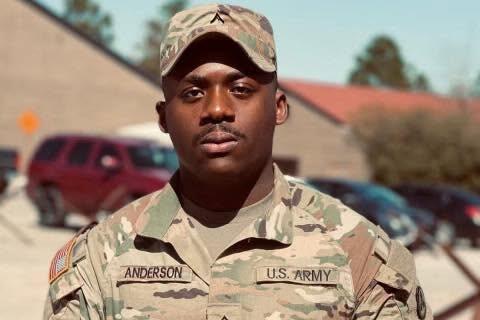 Portrait of a U.S. Army soldier in desert camouflage, cap on, with the name Anderson on the chest, standing outdoors near buildings and cars.