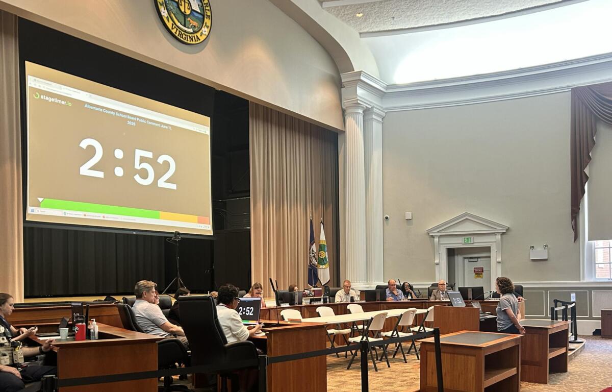 Government meeting in a wood-paneled chamber with officials at desks; a large projector screen shows 2:52 and the Virginia state seal is visible above.