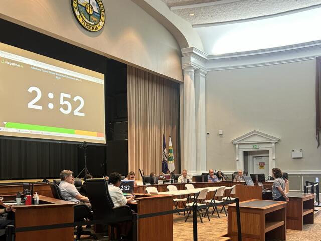 Government meeting in a wood-paneled chamber with officials at desks; a large projector screen shows 2:52 and the Virginia state seal is visible above.
