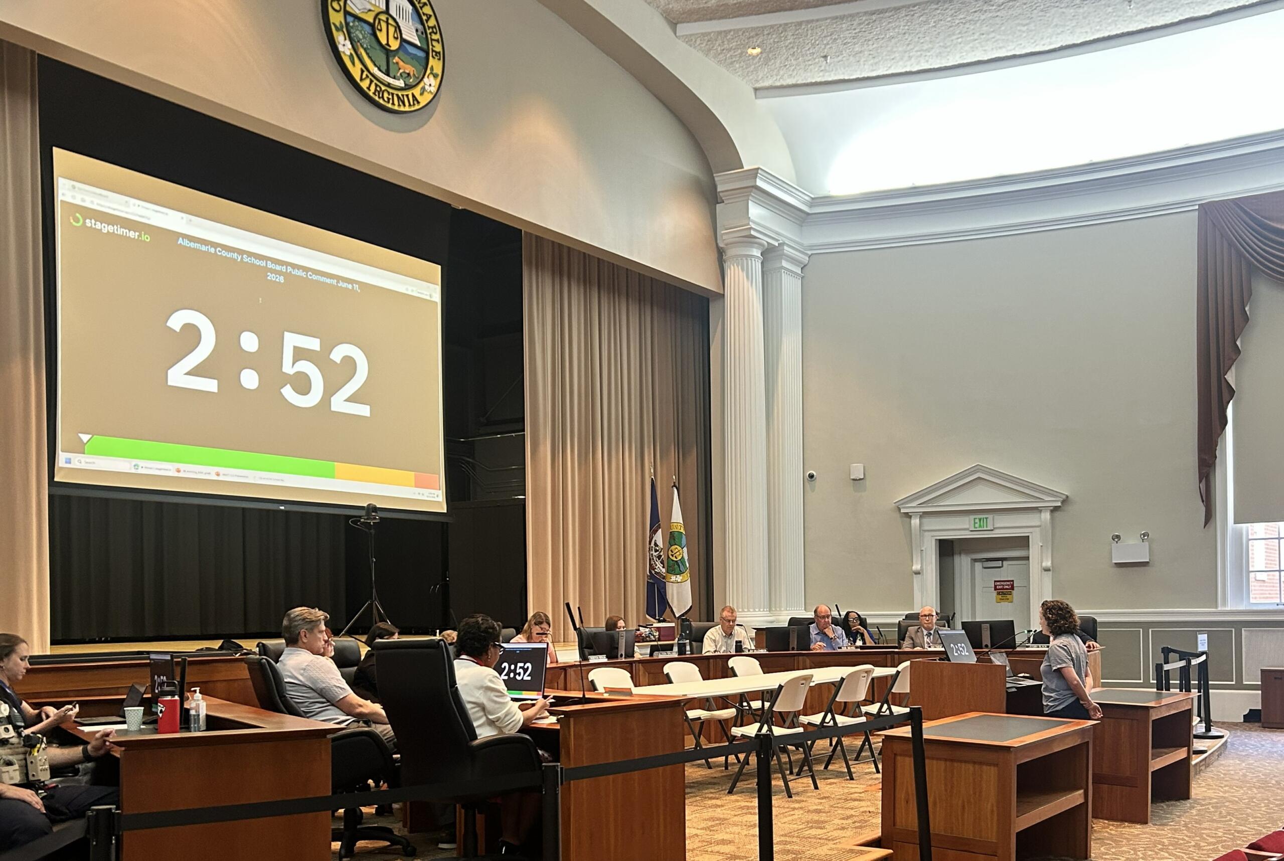 Government meeting in a wood-paneled chamber with officials at desks; a large projector screen shows 2:52 and the Virginia state seal is visible above.