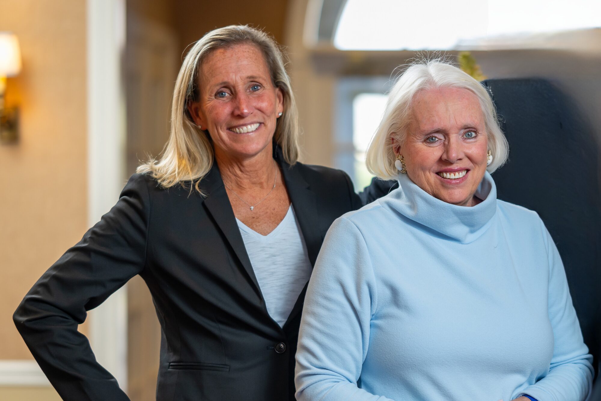 Two smiling women pose together indoors; one in a black blazer and the other in a light blue top.
