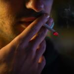 Close-up of a person smoking a cigarette, ember glowing as it rests near their lips in low light.