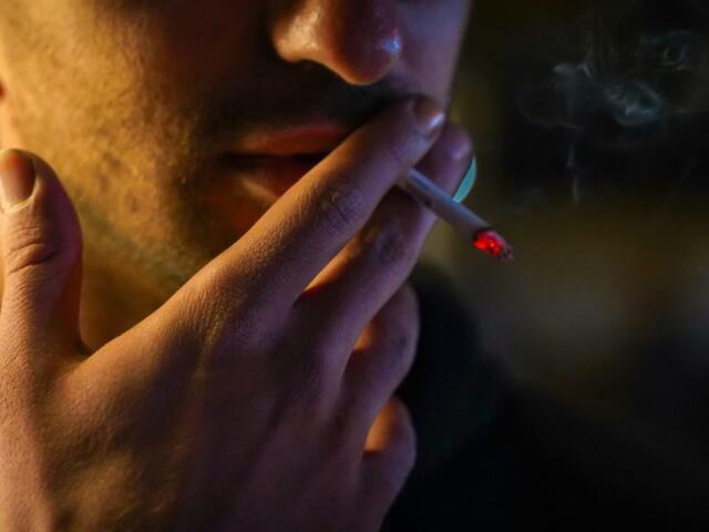 Close-up of a person smoking a cigarette, ember glowing as it rests near their lips in low light.