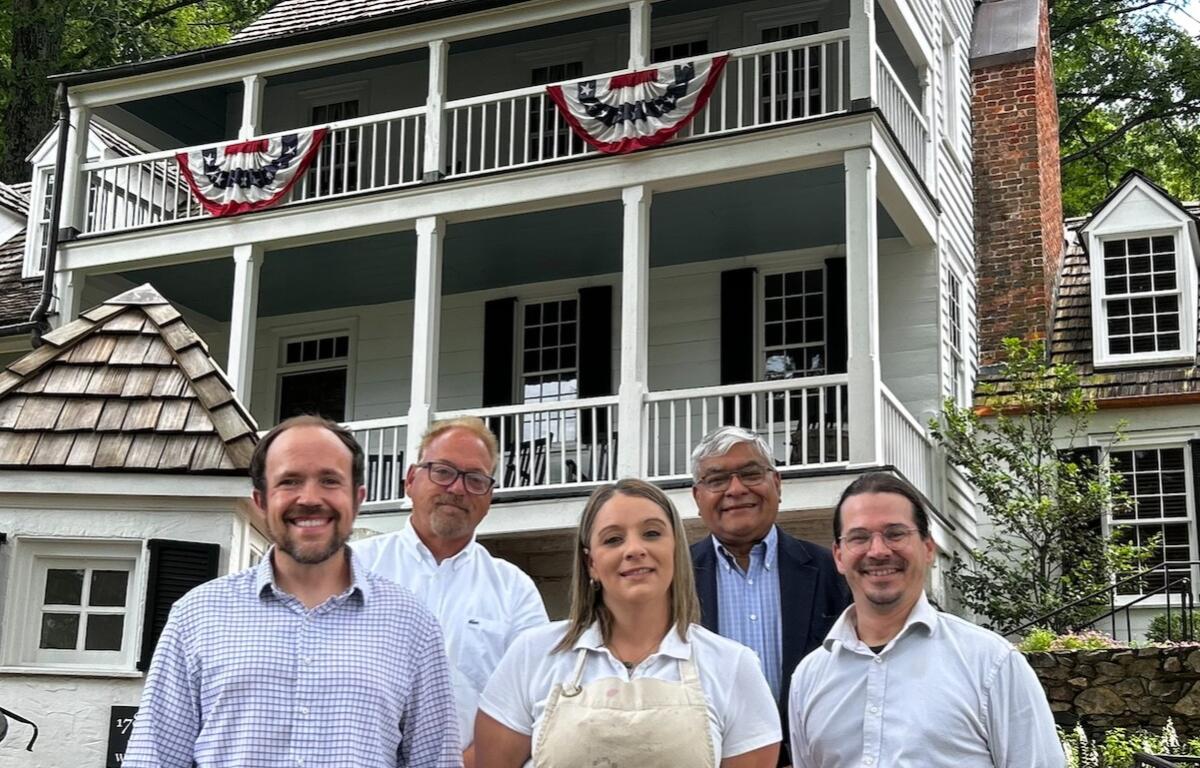Five adults pose in front of a white clapboard house with red, white, and blue bunting on the balcony.
