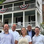 Five adults pose in front of a white clapboard house with red, white, and blue bunting on the balcony.