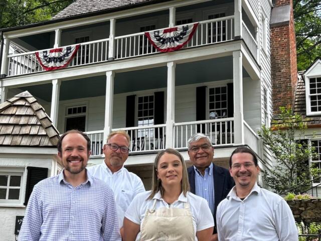 Five adults pose in front of a white clapboard house with red, white, and blue bunting on the balcony.
