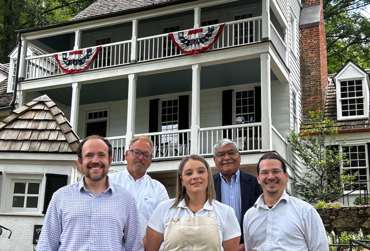 Five adults pose in front of a white clapboard house with red, white, and blue bunting on the balcony.