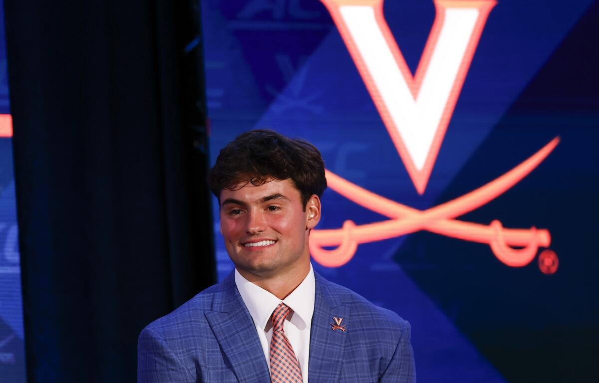 Smiling young man in a blue checkered suit and pink tie on stage, with a Virginia Cavaliers logo backdrop behind him.