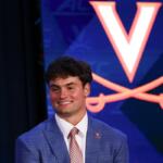 Smiling young man in a blue checkered suit and pink tie on stage, with a Virginia Cavaliers logo backdrop behind him.