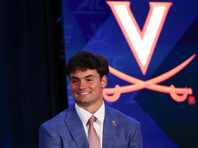 Smiling young man in a blue checkered suit and pink tie on stage, with a Virginia Cavaliers logo backdrop behind him.