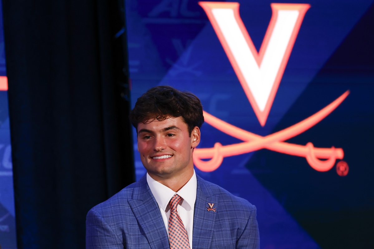 Smiling young man in a blue checkered suit and pink tie on stage, with a Virginia Cavaliers logo backdrop behind him.