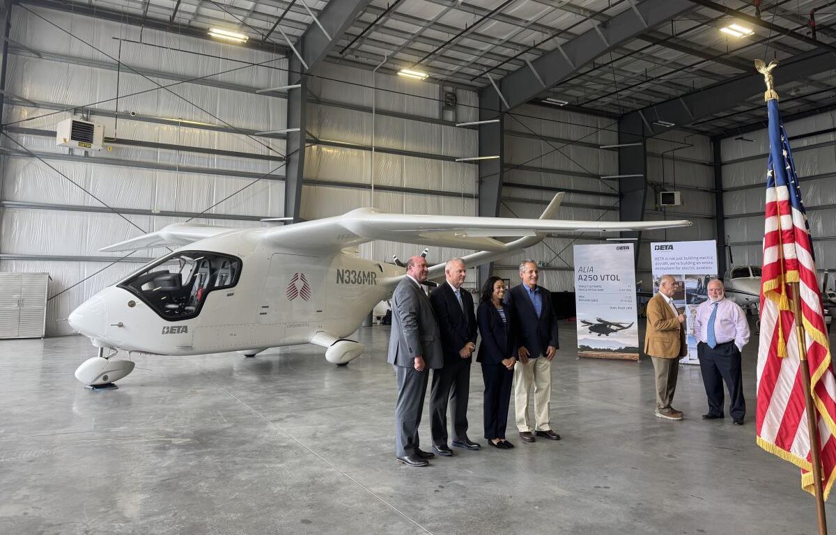 Group of six professionals in suits posing beside a white Beta aircraft inside a hangar, with an American flag in the foreground.
