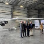 Group of six professionals in suits posing beside a white Beta aircraft inside a hangar, with an American flag in the foreground.