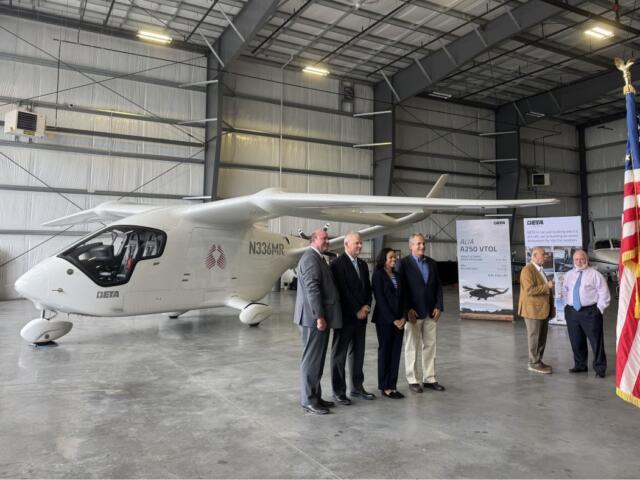 Group of six professionals in suits posing beside a white Beta aircraft inside a hangar, with an American flag in the foreground.
