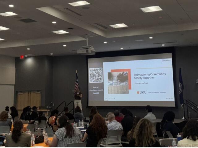 Speaker presenting a slide titled 'Reimagining Community Safety Together' to an audience in a conference room with an American flag nearby