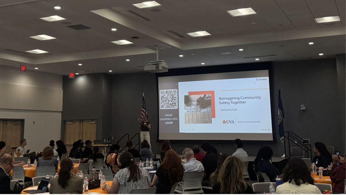 Speaker presenting a slide titled 'Reimagining Community Safety Together' to an audience in a conference room with an American flag nearby