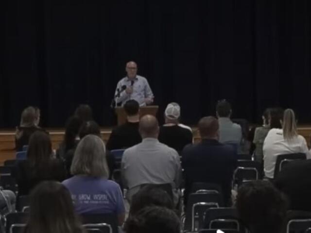 Man at a podium speaking into a microphone as an audience sits facing the stage against a dark curtain backdrop