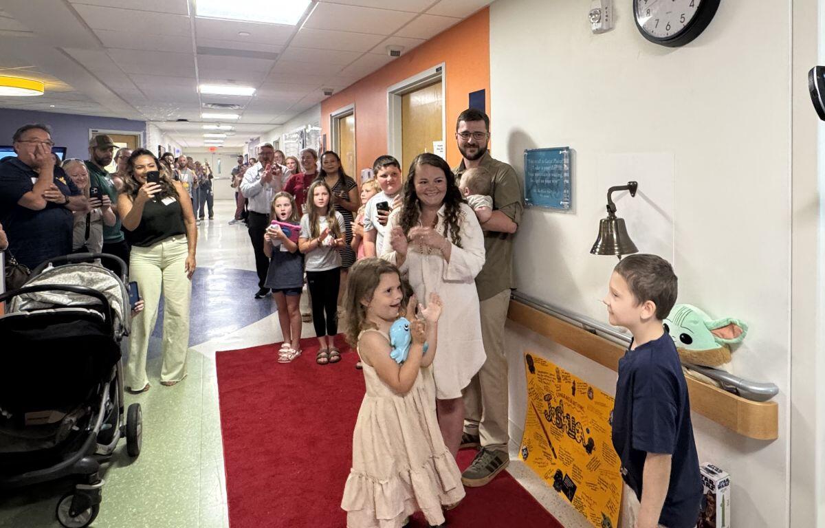 A young girl in a cream dress walks on a red carpet in a hospital hallway as family and staff cheer and take photos.