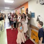 A young girl in a cream dress walks on a red carpet in a hospital hallway as family and staff cheer and take photos.