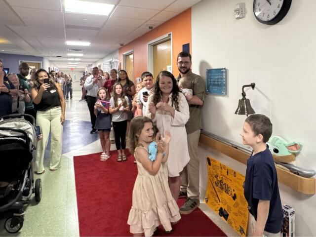 A young girl in a cream dress walks on a red carpet in a hospital hallway as family and staff cheer and take photos.