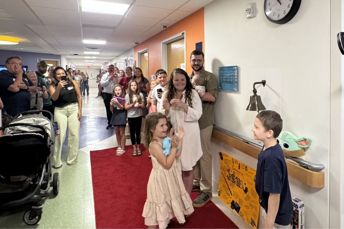 A young girl in a cream dress walks on a red carpet in a hospital hallway as family and staff cheer and take photos.