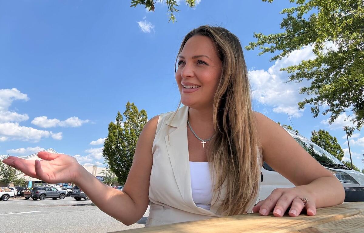 Smiling woman sits at a wooden table outside in a parking lot on a sunny day, gesturing with her left hand. She wears a white top and a light vest with a cross necklace.