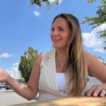 Smiling woman sits at a wooden table outside in a parking lot on a sunny day, gesturing with her left hand. She wears a white top and a light vest with a cross necklace.