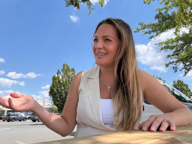 Smiling woman sits at a wooden table outside in a parking lot on a sunny day, gesturing with her left hand. She wears a white top and a light vest with a cross necklace.