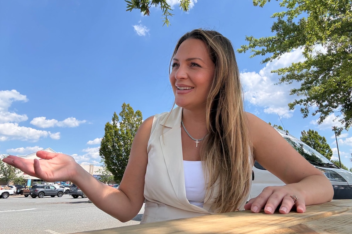 Smiling woman sits at a wooden table outside in a parking lot on a sunny day, gesturing with her left hand. She wears a white top and a light vest with a cross necklace.
