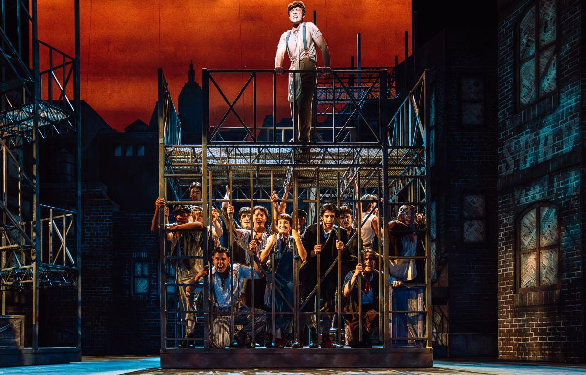 Stage production: a man stands atop a metal cage while a crowd of actors presses against the bars on a city-set, orange sky background.