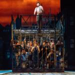 Stage production: a man stands atop a metal cage while a crowd of actors presses against the bars on a city-set, orange sky background.