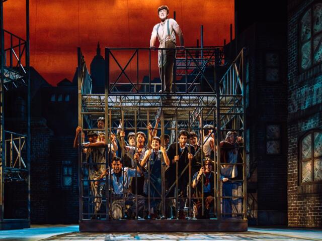 Stage production: a man stands atop a metal cage while a crowd of actors presses against the bars on a city-set, orange sky background.
