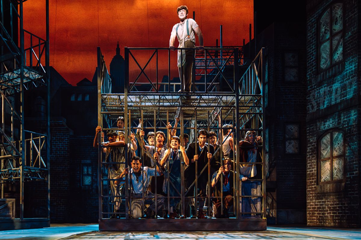 Stage production: a man stands atop a metal cage while a crowd of actors presses against the bars on a city-set, orange sky background.