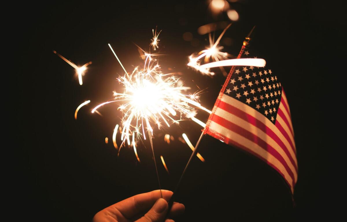 Hand holding a lit sparkler beside a small American flag against a dark background, celebratory mood at night.