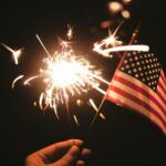 Hand holding a lit sparkler beside a small American flag against a dark background, celebratory mood at night.