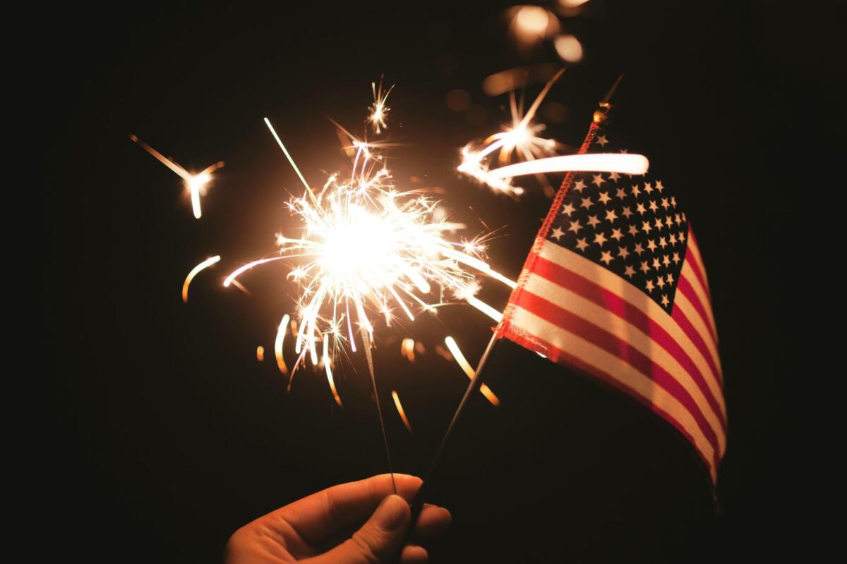 Hand holding a lit sparkler beside a small American flag against a dark background, celebratory mood at night.