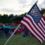 American flag on a pole waved at an outdoor gathering with people blurred in the background