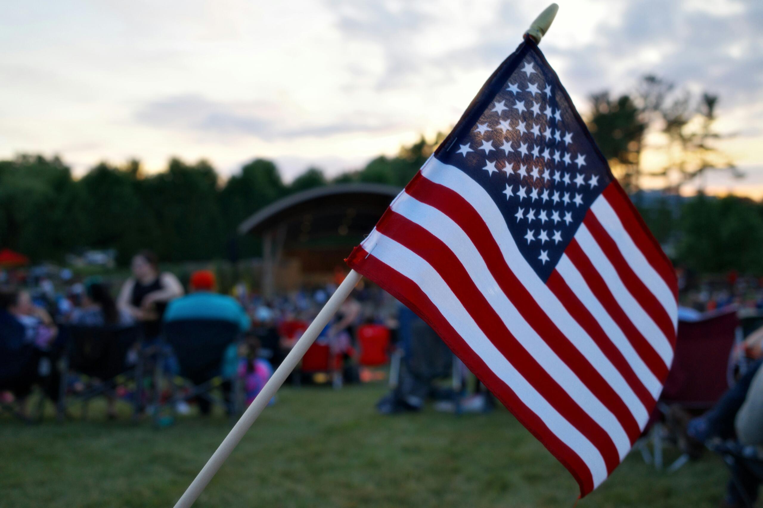 American flag on a pole waved at an outdoor gathering with people blurred in the background