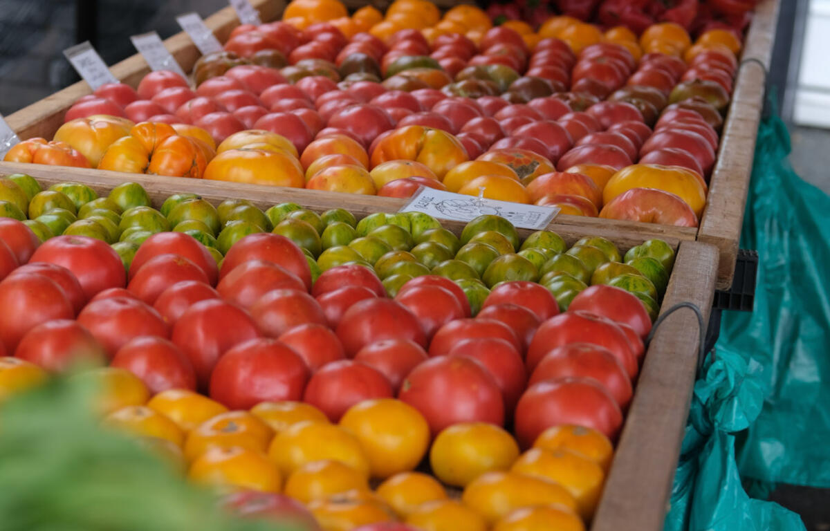 Rows of ripe tomatoes in wooden crates at an outdoor market, with red peppers piled in the back.