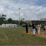 Youth baseball players gather on the infield dirt during practice, with bleachers and a school building in the background on a sunny day.