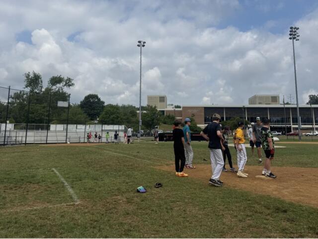 Youth baseball players gather on the infield dirt during practice, with bleachers and a school building in the background on a sunny day.