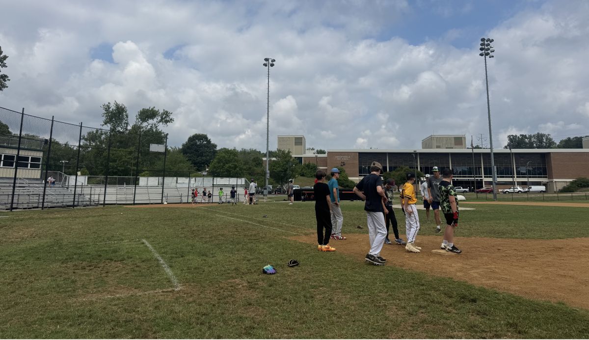 Youth baseball players gather on the infield dirt during practice, with bleachers and a school building in the background on a sunny day.