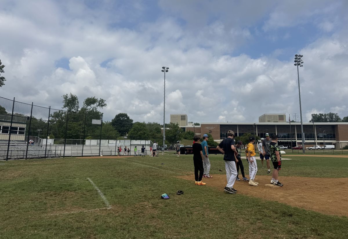 Group of young baseball players standing on the infield dirt during practice, with bleachers and a school building in the background under a cloudy sky.