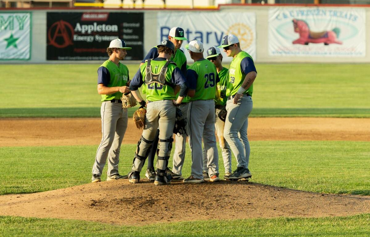 Baseball players in neon vests huddle on the pitcher's mound during a game.