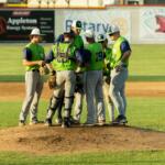 Baseball players in neon vests huddle on the pitcher's mound during a game.