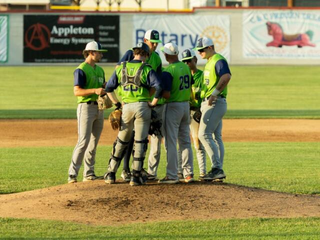 Baseball players in neon vests huddle on the pitcher's mound during a game.