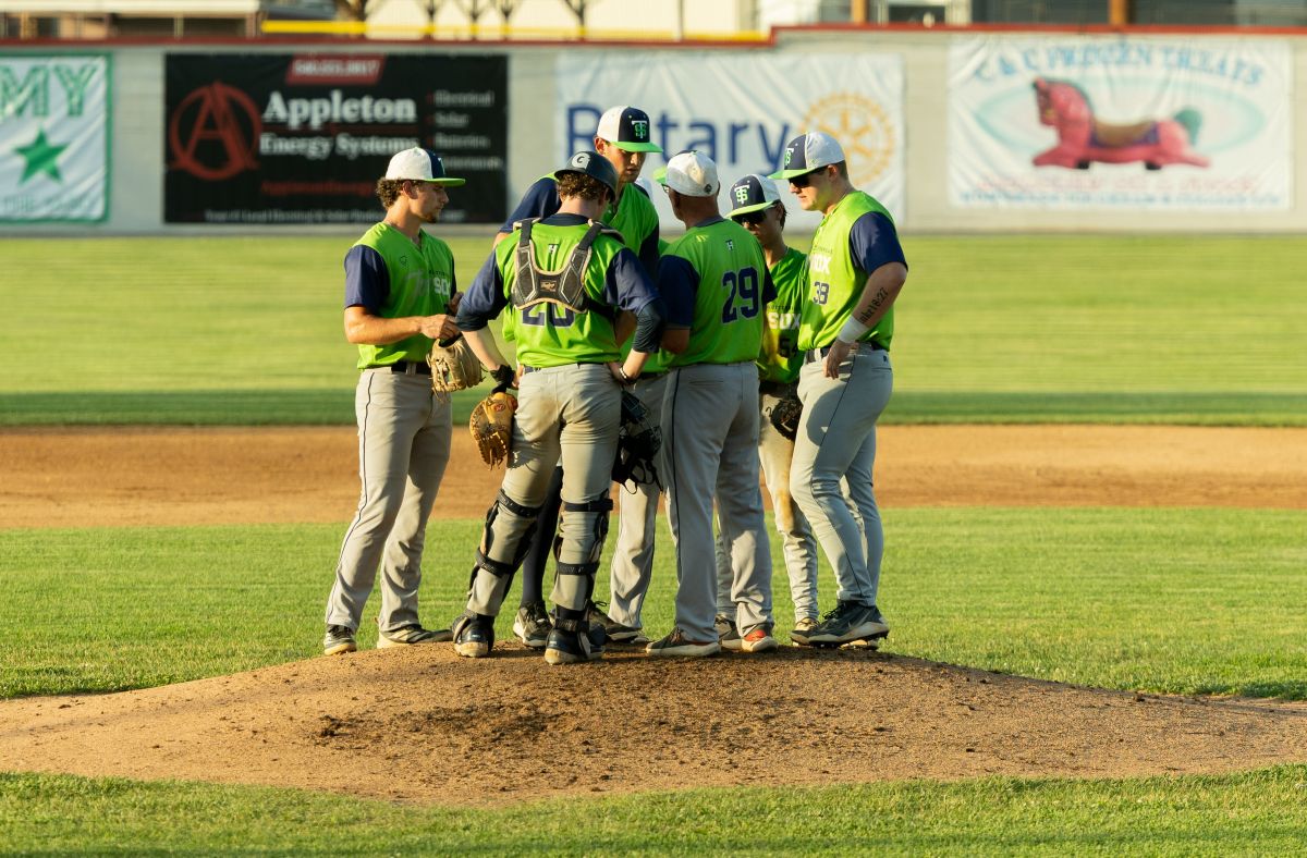 Baseball players in neon vests huddle on the pitcher's mound during a game.
