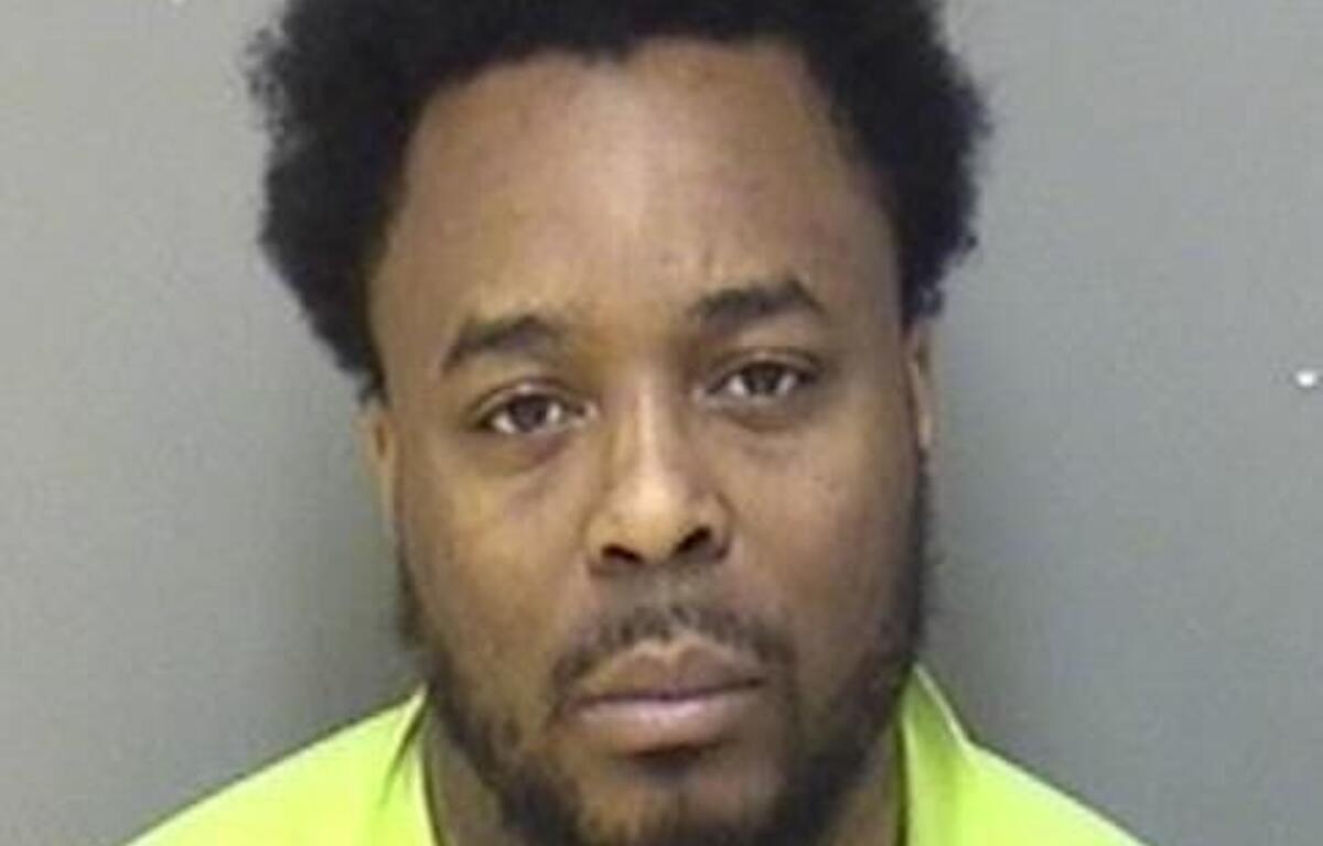 Mugshot of a Black man with short curly hair and beard, wearing a neon yellow safety vest against a gray wall.