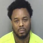 Mugshot of a Black man with short curly hair and beard, wearing a neon yellow safety vest against a gray wall.