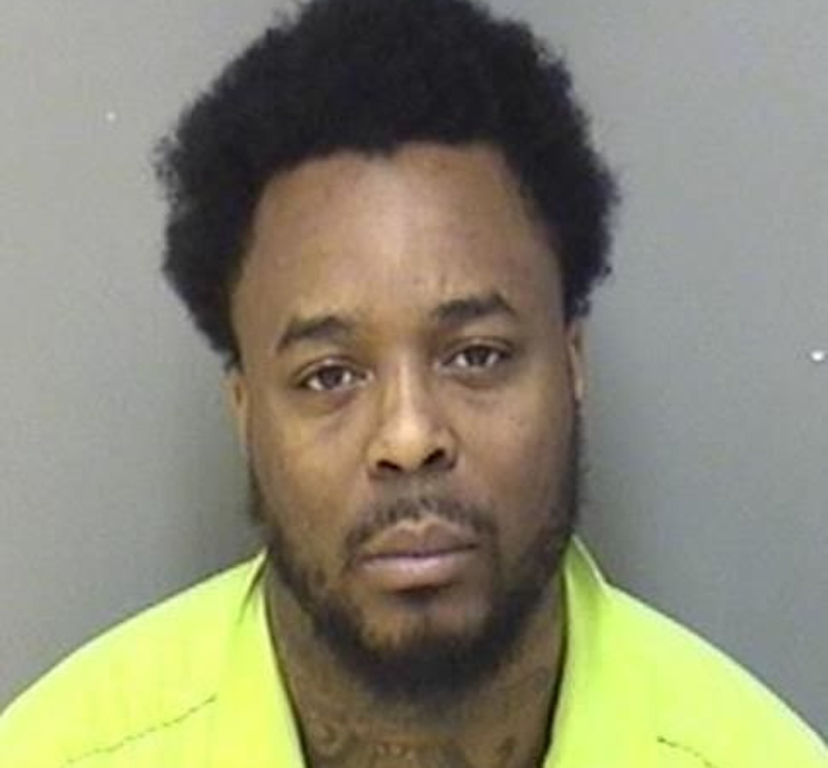 Mugshot of a Black man with short curly hair and beard, wearing a neon yellow safety vest against a gray wall.